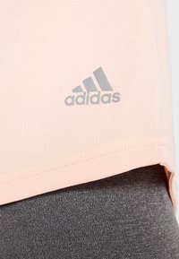 Světle růžové sportovní tričko s texturovanou tkaninou, které má šedé logo Adidas v dolním levém rohu a rovný lem.