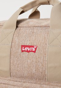 Bolsa de viaje Levi's en tela de color marrón claro. Presenta correas reforzadas en tono marrón y un parche con el logo en rojo. Superficie texturizada y duradera con patrones sutiles.