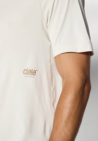 Jasno beżowa koszulka sportowa wykonana z gładkiego materiału, z małym złotym logo "ciele ATHLETICS" po lewej stronie i krótkimi rękawami.