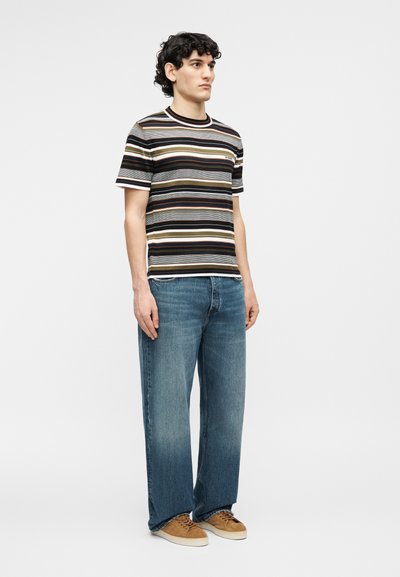 Young man stands wearing a striped short-sleeve shirt, loose blue jeans, and brown sneakers against a plain light background.