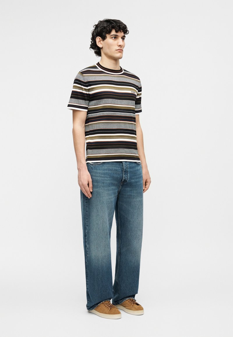 Young man stands wearing a striped short-sleeve shirt, loose blue jeans, and brown sneakers against a plain light background.