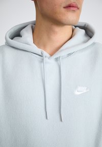 Ljusblå fleecehoodie med en framficka, dragsnoddar vid huvan och en vit Nike-logotyp på bröstet. Mjuk textur.