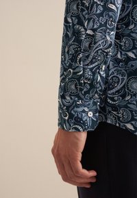 Camicia a maniche lunghe con un motivo paisley blu scuro. Presenta un bottone azzurro a contrasto e una texture di tessuto liscia.