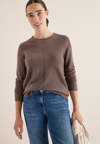 Brauner Strickpullover mit Rundhalsausschnitt, gerippten Bündchen und Saum; kombiniert mit hellblauen Jeans, gehalten mit einem beigen Gegenstand in einer Hand.