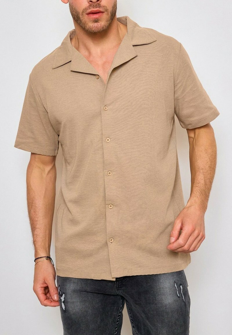 Chemise à manches courtes beige avec un col en V et des boutons sur le devant, fabriquée en tissu texturé. Portée avec un jean foncé déchiré.