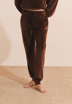 Persona que lleva puestos pantalones jogger marrones de pana con cintura con cordón y puños elásticos, de pie descalza sobre una alfombra beige.