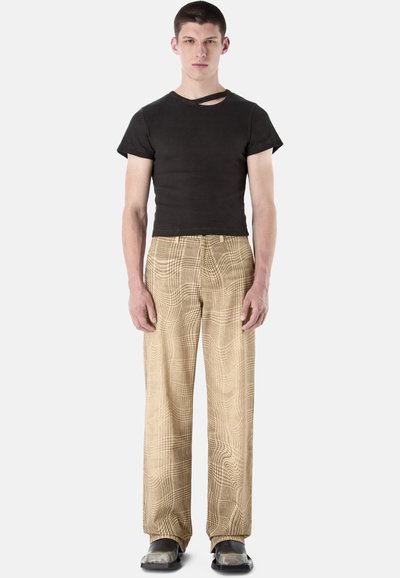 Jeune homme debout face à l'avant, portant un t-shirt noir court avec une découpe et un pantalon large à motifs beige.