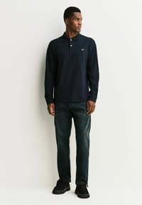 Polo de manga larga en azul marino con tapeta de dos botones y logo en el pecho izquierdo. Se lleva con vaqueros oscuros y calzado negro.