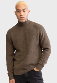 Brown knitted sweater with a high collar, ribbed cuffs, and a textured finish. Worn over dark jeans, featuring a relaxed fit.