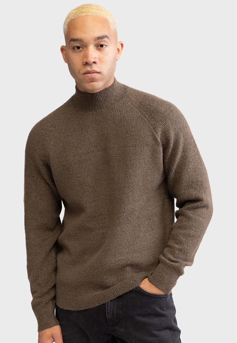 Brown knitted sweater with a high collar, ribbed cuffs, and a textured finish. Worn over dark jeans, featuring a relaxed fit.