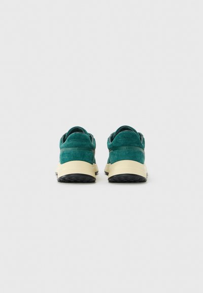 Pair of green suede sneakers with beige midsoles and black outsoles, viewed from the back on a white background.