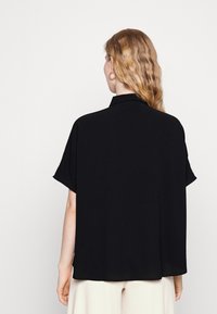 Svart kortärmad skjorta med avslappnad passform, utrustad med en platt krage och slät textur. Bakifrån syns en lös, flödande design.