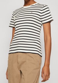 Pruhované tričko v krémové a námořnické modré barvě, krátké rukávy, kulatý výstřih, s malým logem na hrudi. V kombinaci s béžovými kalhotami.