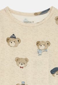 Svijetlo bež majica s različitim grafičkim prikazima medvjeda koji nose šešire i dodatke. Izrađena od mekog materijala s okruglim izrezom i tiskanim uzorkom.