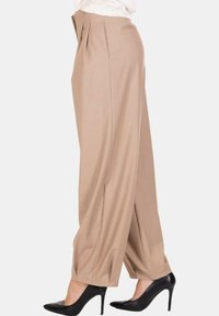 Pantaloni beige a gamba larga indossati con scarpe nere con tacco alto, mostrati di profilo su uno sfondo bianco semplice.