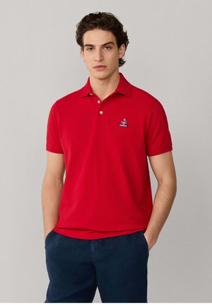Joven con polo rojo de manga corta con un pequeño emblema bordado, de pie con las manos en los bolsillos de unos pantalones azul marino.