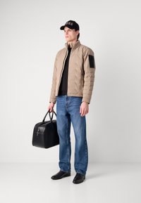 Beige puffjacka med ståkrage och svart axelpanel, i kombination med svart skjorta och blå jeans. Håller en svart läderväska och har på sig svarta skor.