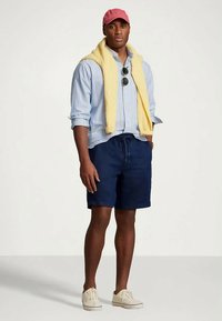 Błękitna koszula w paski, żółty sweter zawiązany na szyi, granatowe szorty, różowa czapka z daszkiem, okulary przeciwsłoneczne i białe buty. Codzienny, letni strój.