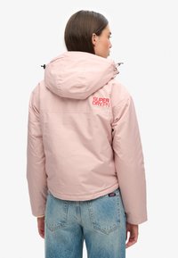 Różowa kurtka z kapturem o błyszczącej powierzchni, z czerwoną logo "Superdry" z tyłu. Krótki krój połączony z jasnoniebieskimi dżinsami.