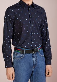 Man som bär en marinblå skjorta med ljusblå prickar, blåjeans och ett svart bälte med regnbågsränder.