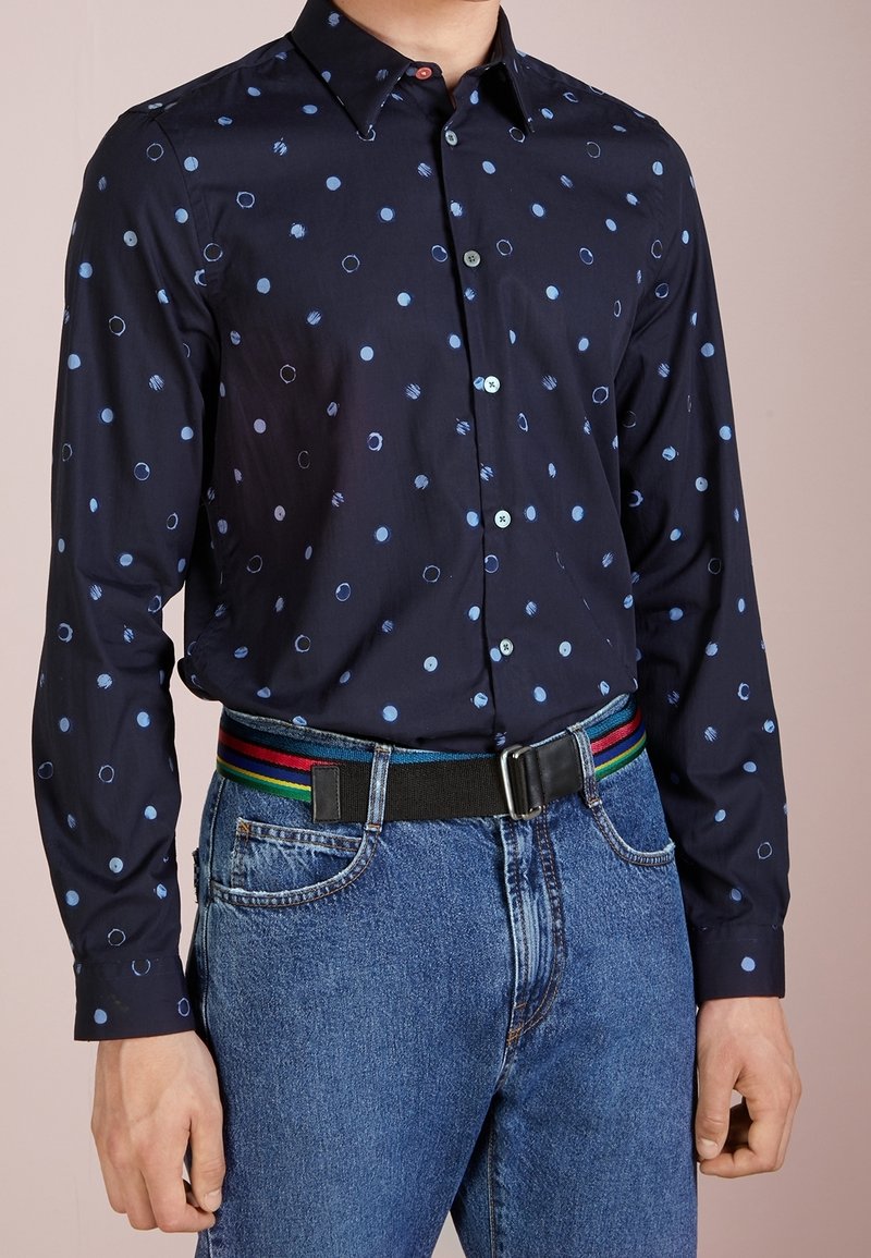 Man som bär en marinblå skjorta med ljusblå prickar, blåjeans och ett svart bälte med regnbågsränder.