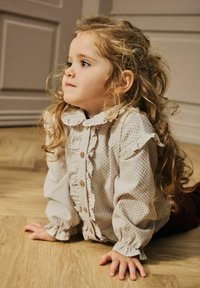 Blusa beige chiaro con pois, con colletto e polsini arricciati, caratterizzata da bottoni in legno. Modella in ginocchio su un pavimento di legno.