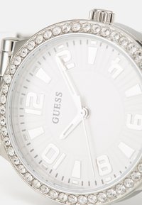 Orologio argento con un bezel accentato di cristalli, quadrante bianco, indicatori delle ore audaci e lancette argento, con il nome del marchio "GUESS" ben in evidenza.