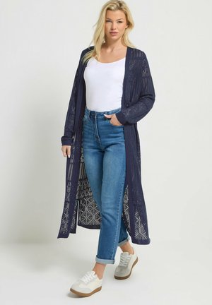 Lyshåret kvinde iført en lang marineblå strikket cardigan, hvid tanktop, blå op-rullede jeans og hvide sneakers, stående foran en ensfarvet baggrund.