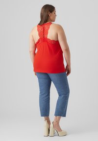Red sleeveless top with lace back detail, paired with high-waisted blue jeans and beige heeled shoes. Plain gray background.