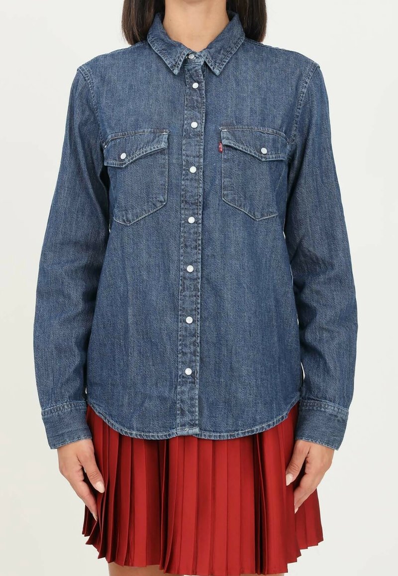 Femme portant une chemise en jean bleue à boutons avec poches poitrine et une jupe plissée rouge, debout devant un fond clair uni.