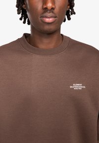 Sudadera marrón hecha de material suave, con cuello redondo y un logo blanco en el lado izquierdo del pecho. Diseño sencillo sin patrones ni acentos.