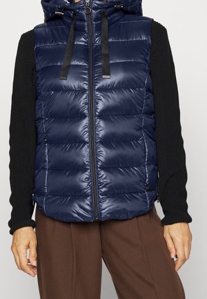 Personne portant une doudoune sans manches matelassée bleu marine avec capuche et manches en maille noire, assortie à un pantalon marron.
