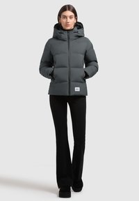 Chaqueta de plumas gris con capucha, cierre de cremallera y parche de logo. Cuenta con un diseño acolchado y bolsillos laterales. Combinada con pantalones y zapatos negros.