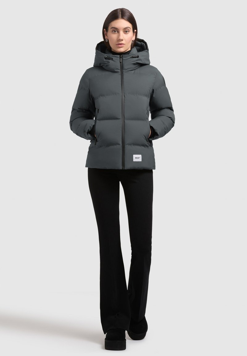 Chaqueta de plumas gris con capucha, cierre de cremallera y parche de logo. Cuenta con un diseño acolchado y bolsillos laterales. Combinada con pantalones y zapatos negros.