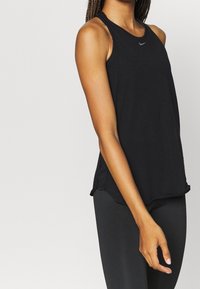 Kvinna i en svart ärmlös Nike-träningsopp och svarta leggings, stående mot en enkel ljus bakgrund.