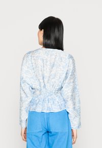 Blusa azul clara com um tecido texturizado e padrão de nuvens, com cintura franzida e mangas longas. Usada com calças azuis.