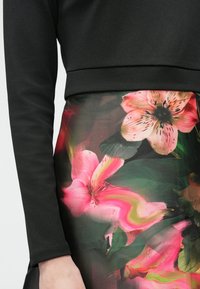 Kadar blizu crnog dugog rukava koji je uparen s suknjom s velikim ružičastim i zelenim cvjetnim uzorkom na tamnoj pozadini.