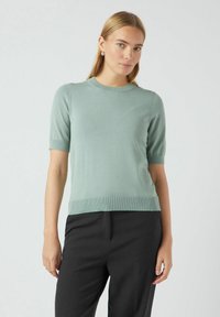 Svijetlozelena kratkoperjana pletena majica s okruglim izrezom, rebrastim rubom i glatkom teksturom, u kombinaciji s crnim hlačama ravnog kroja.