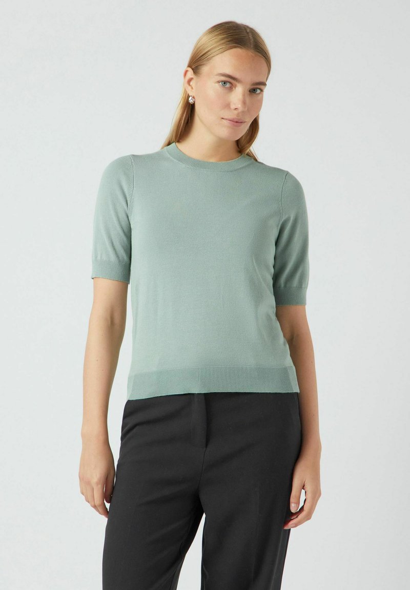 Svijetlozelena kratkoperjana pletena majica s okruglim izrezom, rebrastim rubom i glatkom teksturom, u kombinaciji s crnim hlačama ravnog kroja.