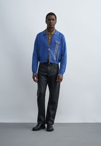 Homme debout portant une veste bleu zippée sur un polo marron, un pantalon en cuir noir et des chaussures noires sur un fond gris clair.