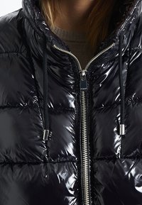 Lesklá černá péřová bunda s vysokým límcem, zipovým zapínáním a stahovacími šňůrkami. Obsahuje prošívané části a stříbrné kovové doplňky.