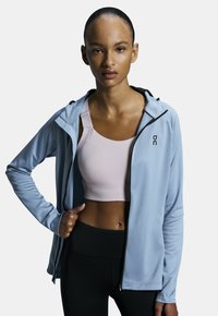 Ljusblå zip-jacka för träning med huva, bärs över en rosa sport-bh. Tyget verkar slätt med en subtil textur. Svarta accenter är närvarande.