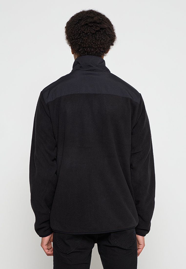 Veste en polaire noire avec un col haut, un panneau arrière texturé et des manches longues. Design minimaliste sans marque ni motifs visibles.