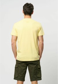 Tricou scurt galben cu mânecă scurtă, dintr-un material texturat, având un guler rotund și o printare subtilă a logo-ului pe partea superioară a spatelui. Asociat cu șorturi cargo de culoare măslinie.
