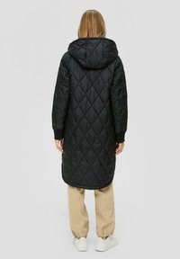 Schwarzer, gesteppter Langmantel mit Kapuze, Rautenmuster und gerippten Bündchen, kombiniert mit beigen Hosen und weißen Sneakers. Glatte, leichte Textur.
