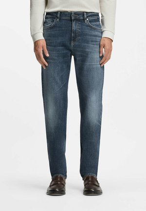 Mand iført falmede blå straight-leg jeans med mørkebrune loafers og en lyse farvet langærmet skjorte, står foran en hvid baggrund.