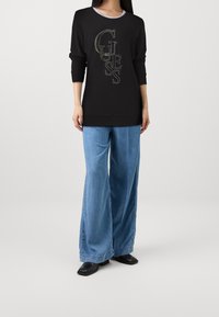 Pull noir avec logo "GUESS" en fil métallique, associé à un jean en denim bleu clair à jambes larges et des bottines noires. Tissu doux, coupe décontractée.