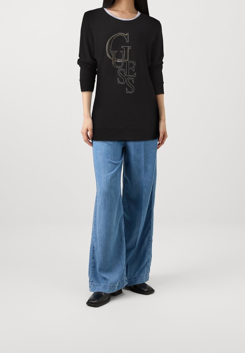 Pull noir avec logo "GUESS" en fil métallique, associé à un jean en denim bleu clair à jambes larges et des bottines noires. Tissu doux, coupe décontractée.