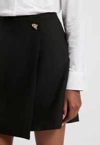 Gonna nera con dettaglio di una spilla a forma di cuore dorata. Realizzata in tessuto liscio, presenta un design avvolgente e una finitura pulita. Abbinata a una camicia bianca.