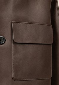 Manteau en cuir marron en gros plan montrant une grande poche rectangulaire avant avec rabat et un bouton noir à quatre trous sur le côté.